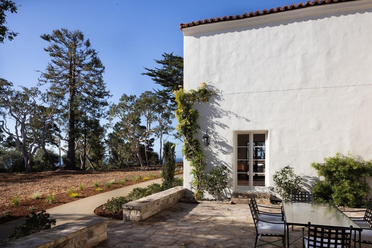 White house with a patio and a table, surrounded by trees and a path leading to a nice view.