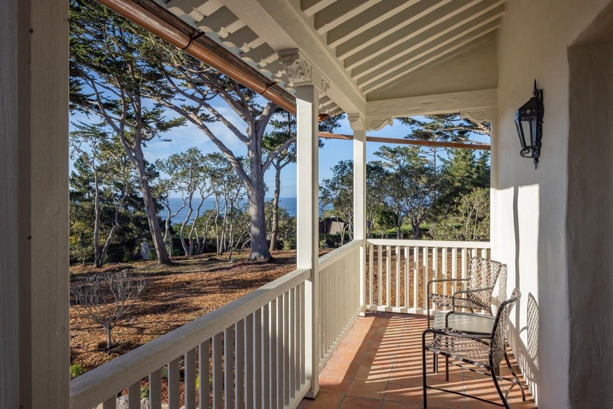 Sun-dappled veranda with a white railing and a pair of chairs, offering a picturesque view of a verdant landscape and the distant ocean.