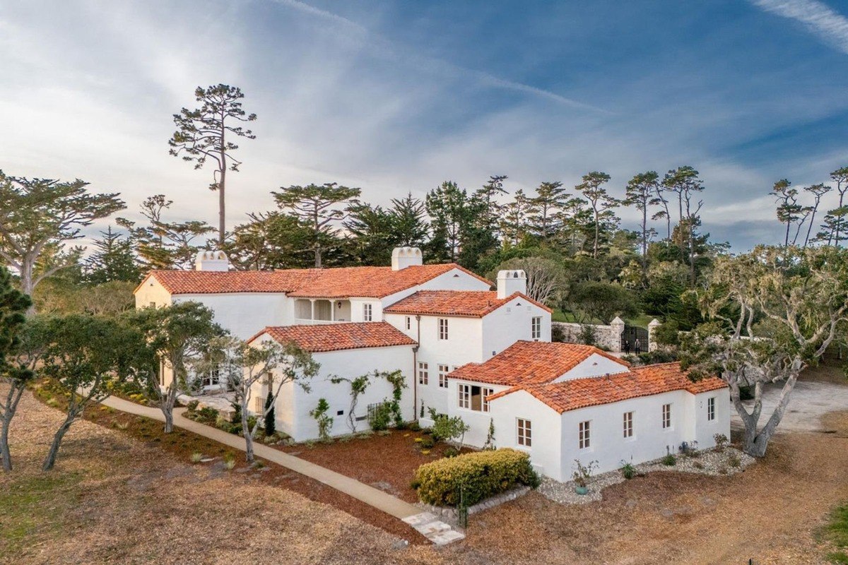 Aerial view of a sprawling estate at dusk, showcasing a white stucco main residence with a terracotta roof, surrounded by mature trees and illuminated pathways.