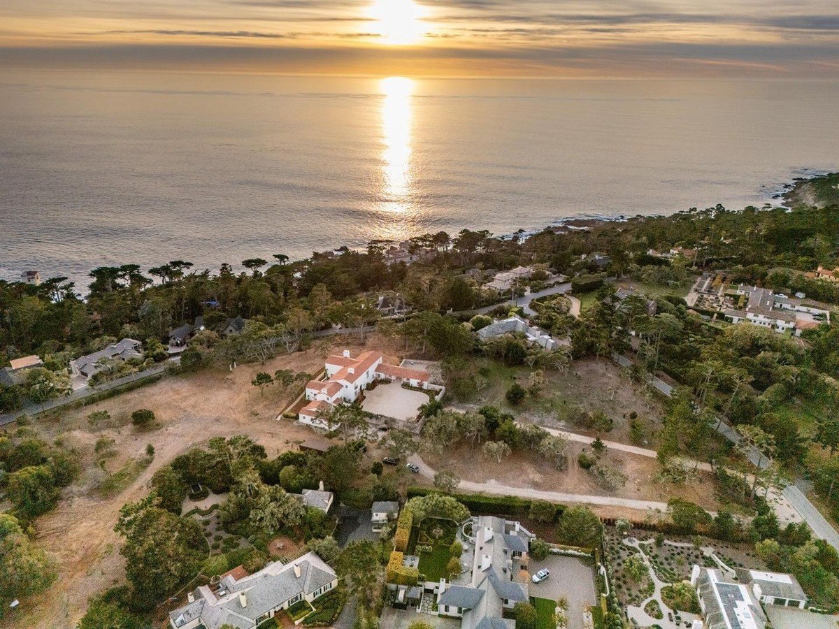 Panoramic aerial view captures a picturesque coastal estate at sunset, showcasing a sprawling property with lush greenery, a white stucco residence, and the sun casting a radiant glow over the Pacific Ocean.