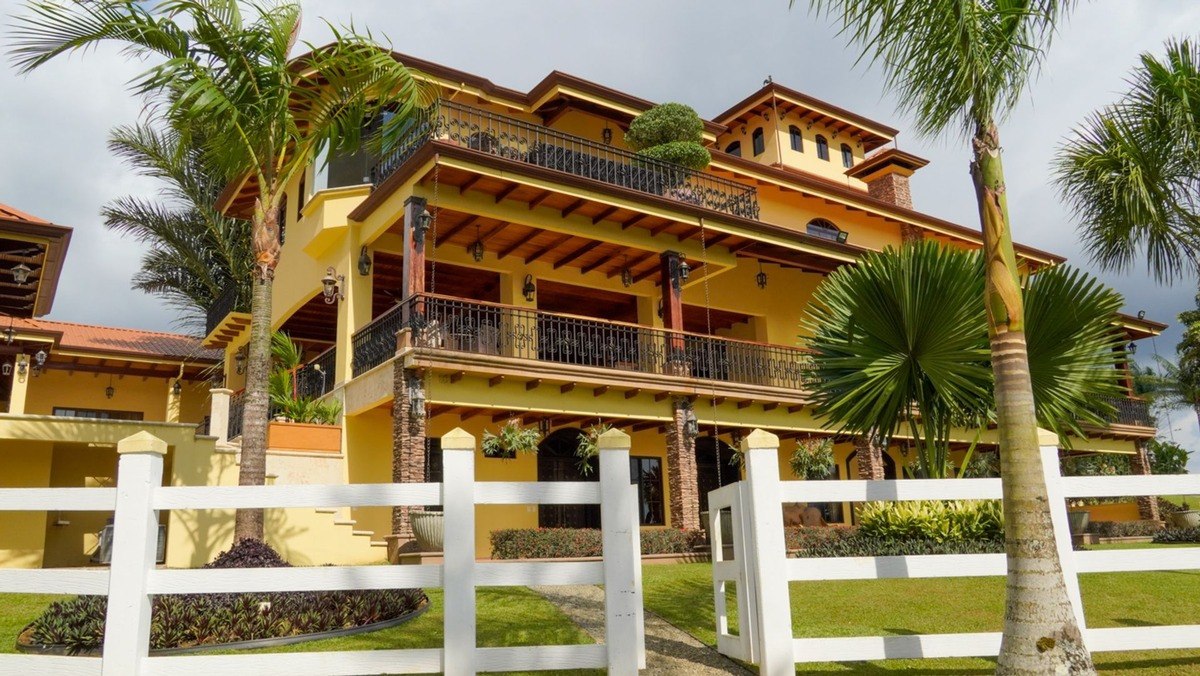 Large yellow house has multiple balconies and tropical landscaping.