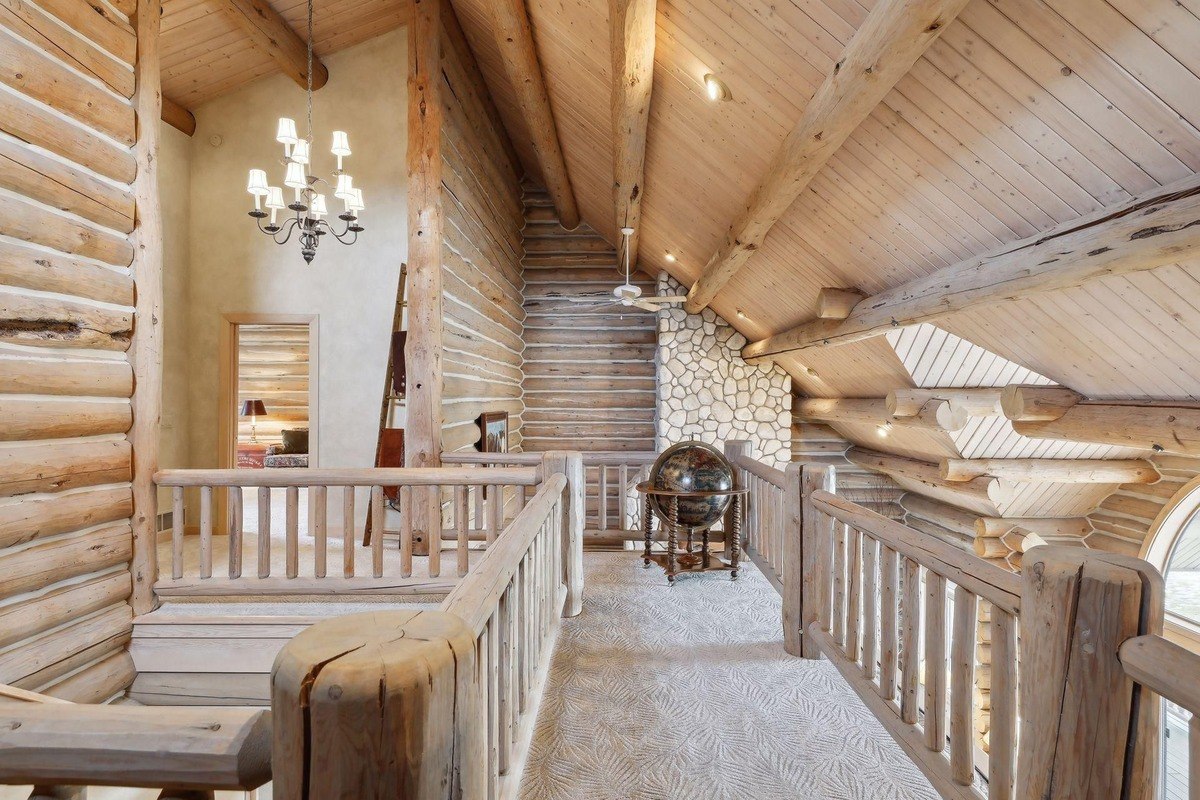 An interior view of a rustic log cabin loft with natural wood beams, railings, and walls, a vaulted ceiling, a stone accent wall, a chandelier, and a decorative globe on a wooden stand, with carpeted flooring featuring