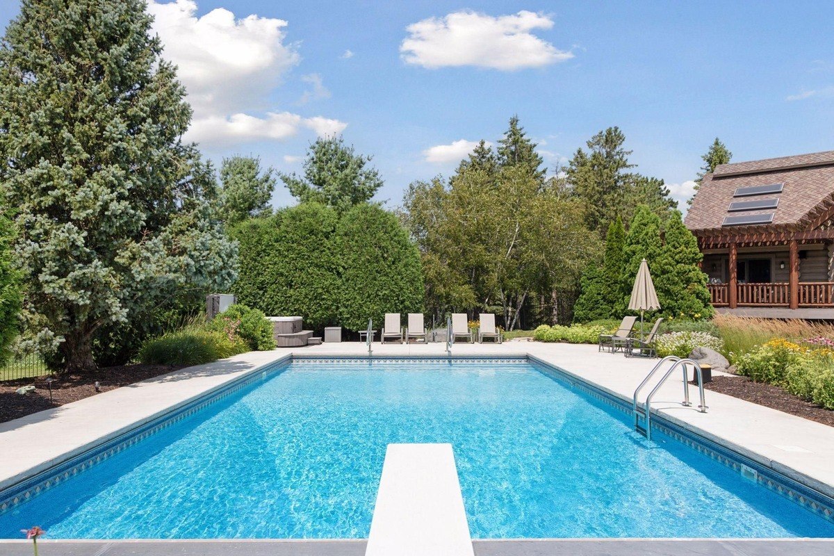 Backyard swimming pool surrounded by landscaping, patio furniture, and tall trees.