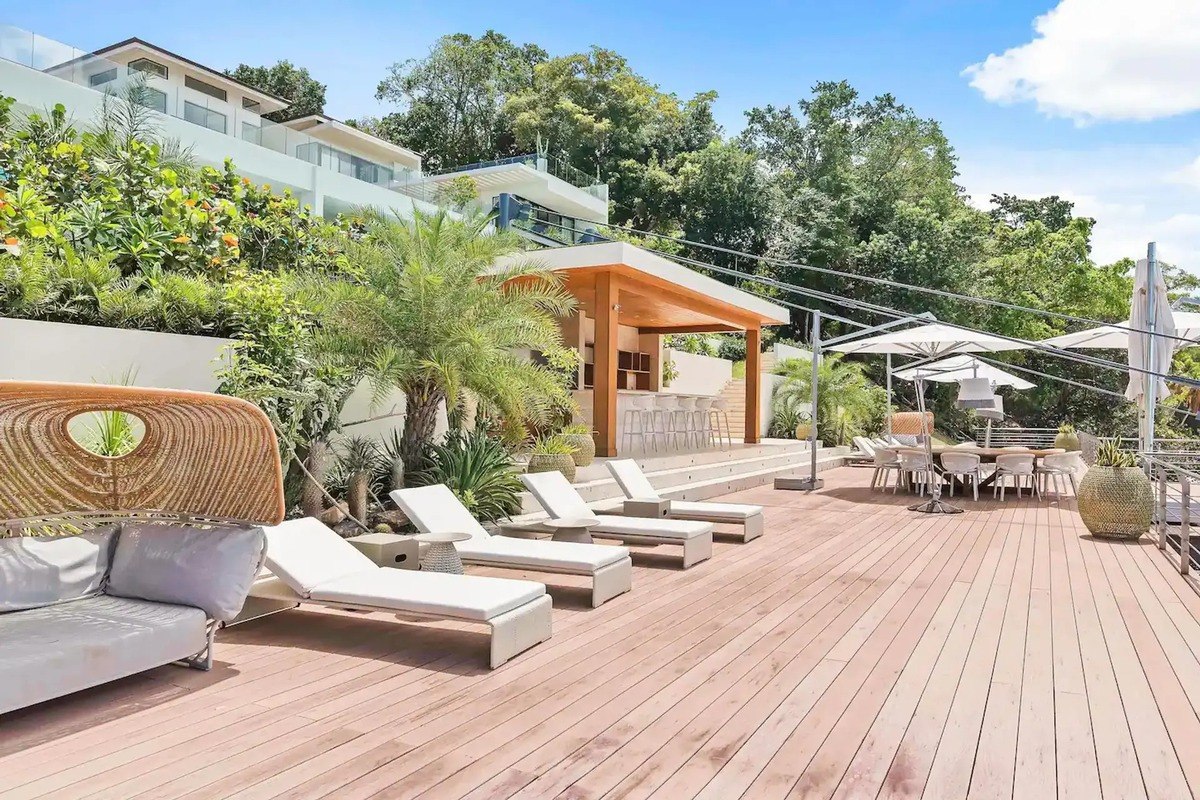 Large deck has lounge chairs, outdoor bar, and large home in background.