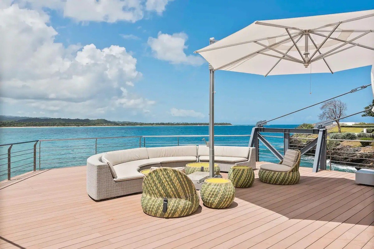 Oceanfront deck has curved sectional sofa, umbrella, and woven chairs.