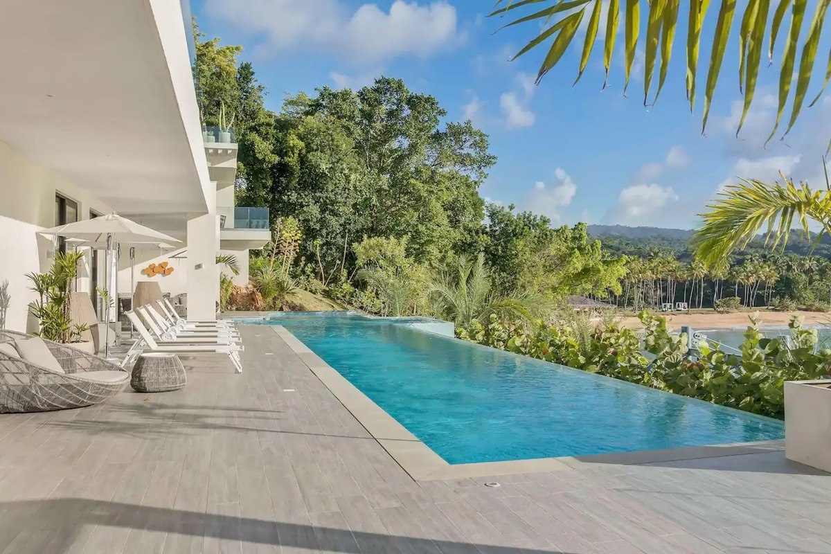 Infinity pool has lounge chairs, umbrella, and tropical view.