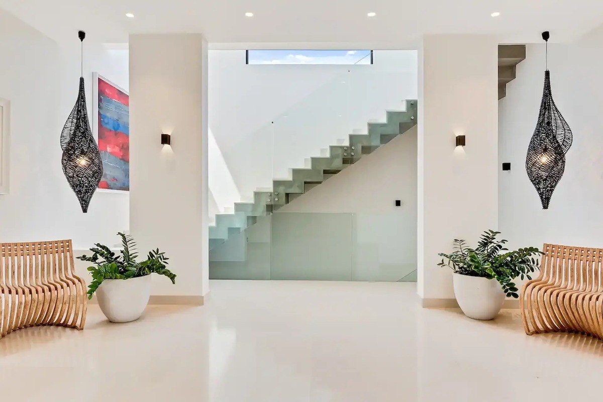 Modern foyer has glass staircase, light wood benches, and hanging light fixtures.