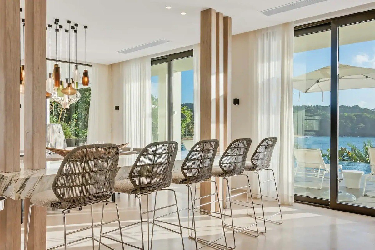 Dining area has woven bar stools, large windows overlooking water, and modern light fixture.