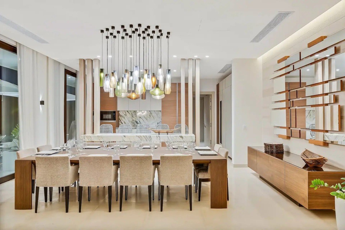 Dining room has large wooden table with beige chairs, modern light fixture, and wood wall decor.