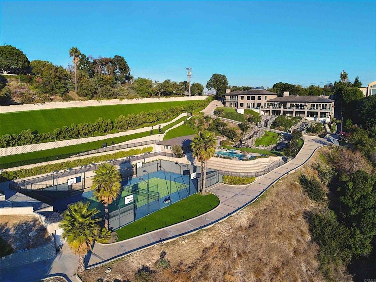 Elevated view of a large estate featuring a tennis court, landscaped paths, and multi-level architecture surrounded by lush greenery.