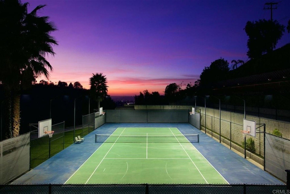 Tennis court illuminated against a vibrant dusk sky, showcasing built-in basketball hoops and seating.