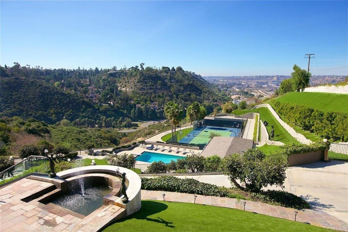 Elevated perspective showcasing a landscaped hill, a pool, and a tennis court framed by greenery.