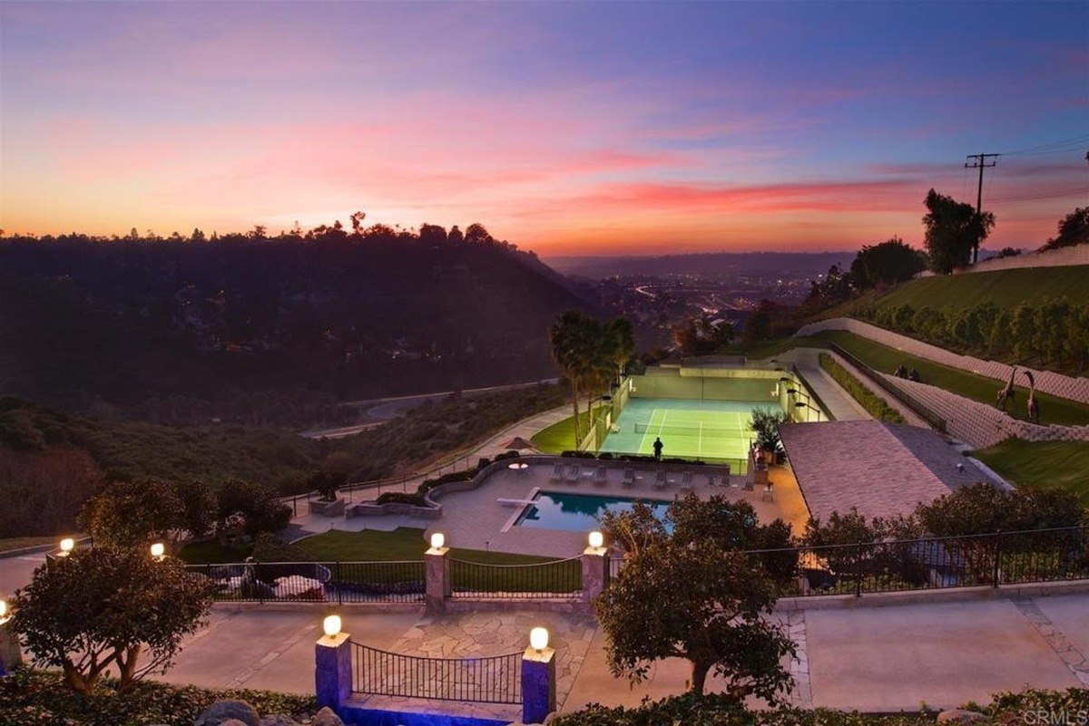 Evening view features a glowing tennis court, a pool, and vibrant landscaping against a vivid sunset backdrop.