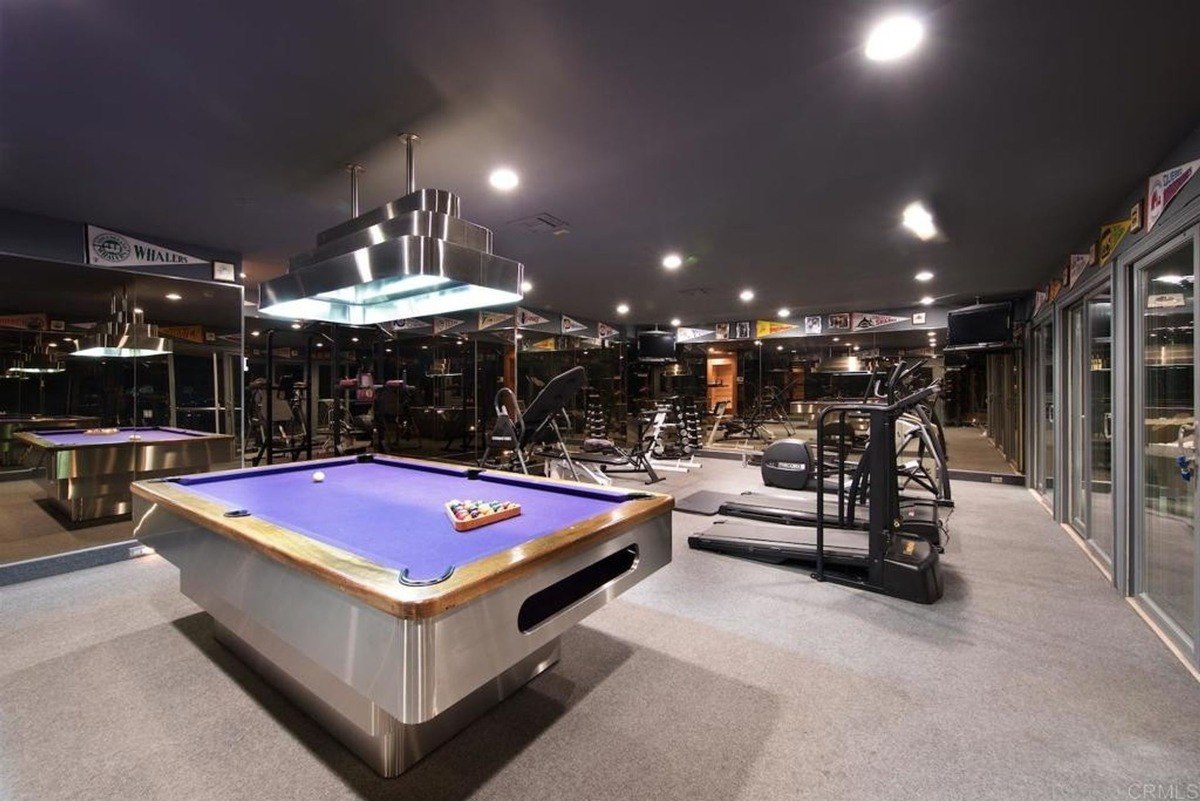 Recreation and fitness area combining pool tables and exercise equipment, illuminated by pendant lights.