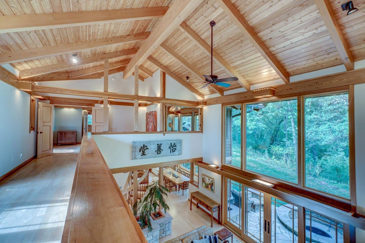 Second-story overlook reveals a spacious open-concept living area with a vaulted ceiling, exposed beams, and large windows offering views of the surrounding landscape.