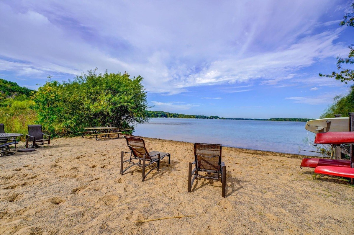 Lounge chairs and a picnic table invite relaxation on a sandy beach with a view of the calm lake and a blue sky.