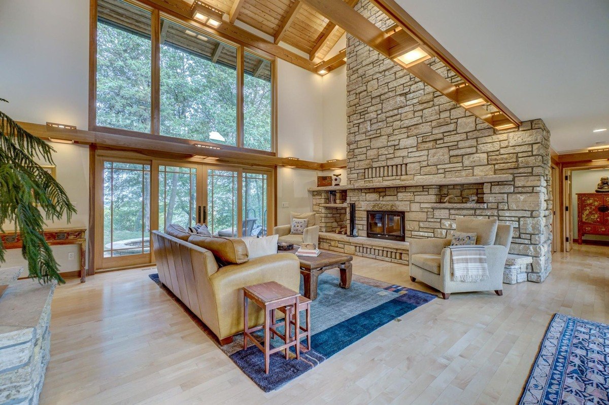 Spacious living room with a soaring ceiling and large windows showcases a cozy seating arrangement around a stone fireplace.