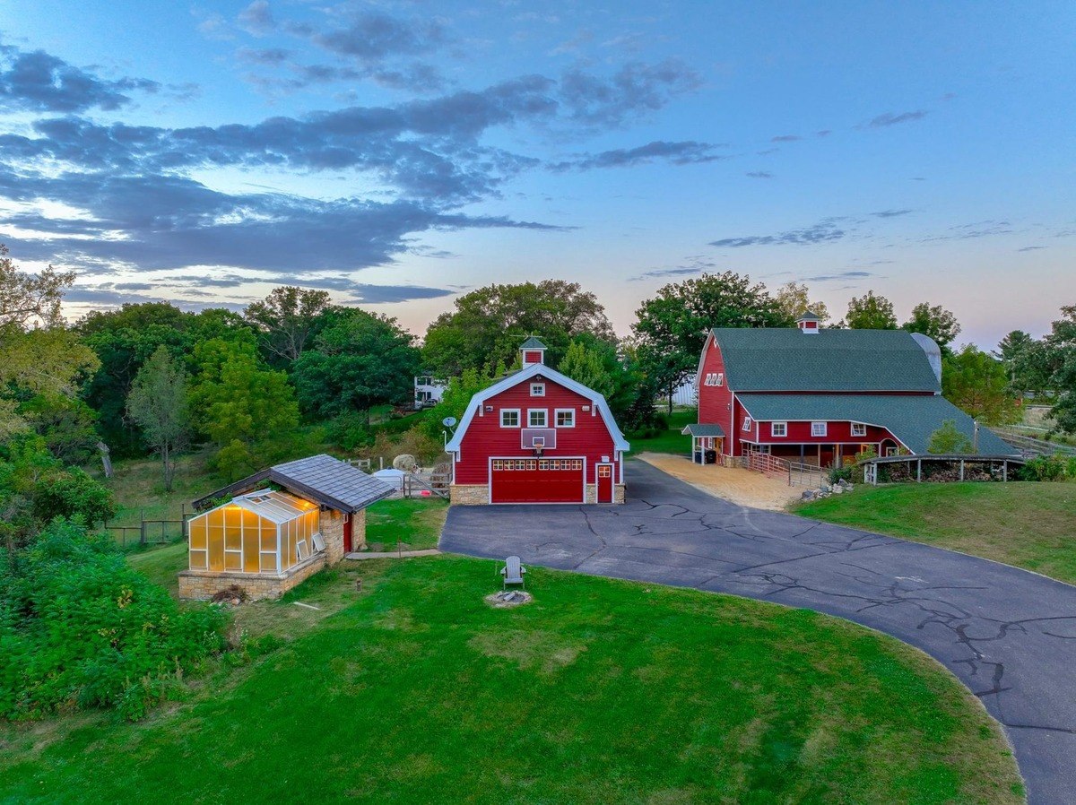 Picturesque rural scene features two red barns, a greenhouse, and a driveway winding through a lush green landscape under a vibrant evening sky.