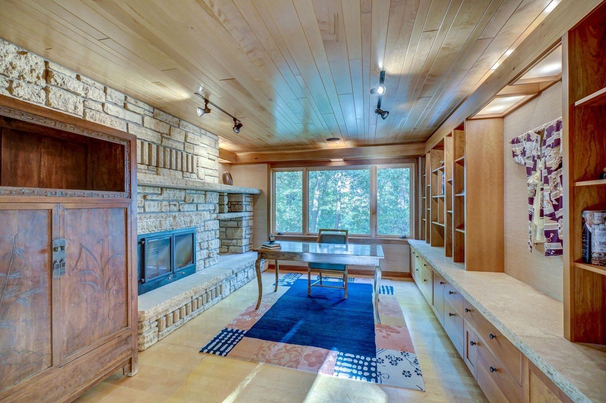 Home office with a wood-paneled ceiling and stone fireplace offers a tranquil workspace with built-in shelving and a view of the outdoors.