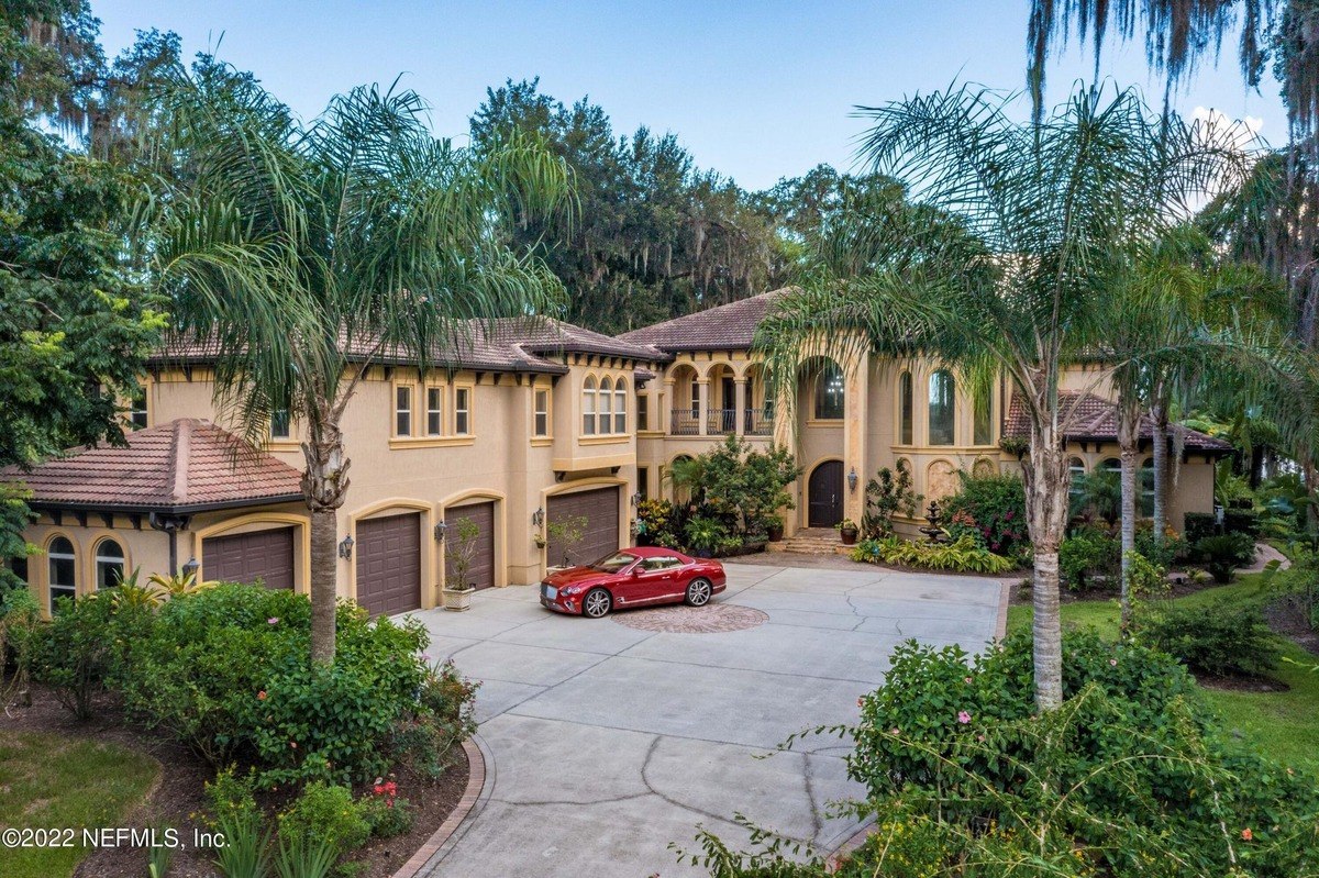 Large, tan house with a red car parked in front, surrounded by palm trees and landscaping.