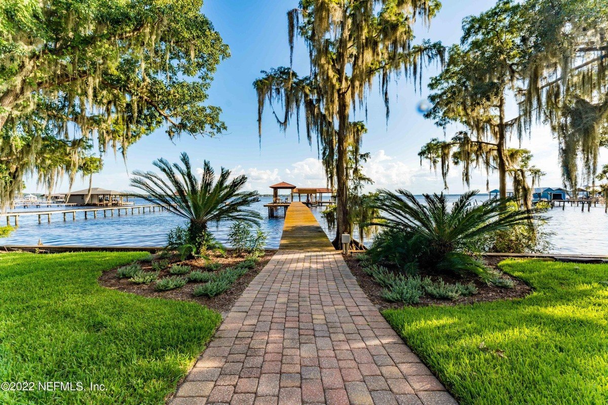 Brick walkway leads to a dock on a lake with lush trees.