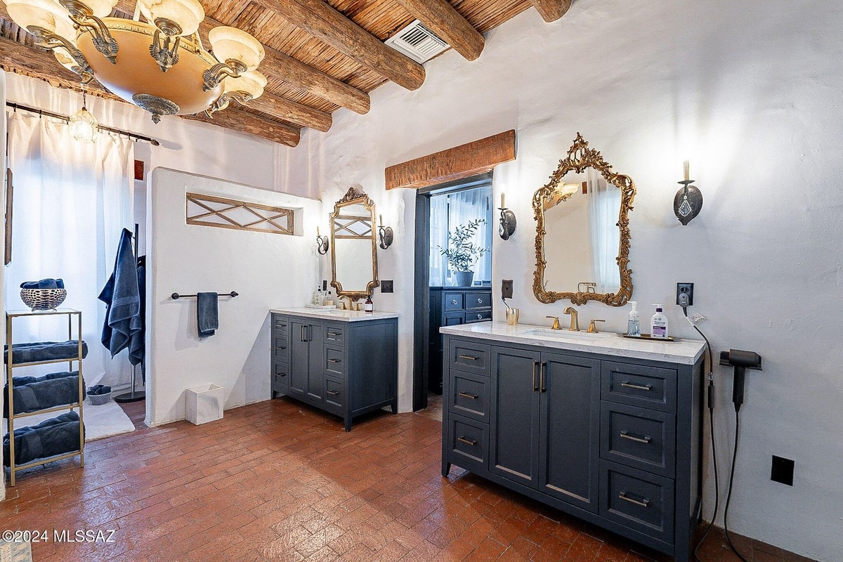 Large bathroom with terracotta tile floors, exposed wood beams, and two dark gray double vanities with ornate gold mirrors.