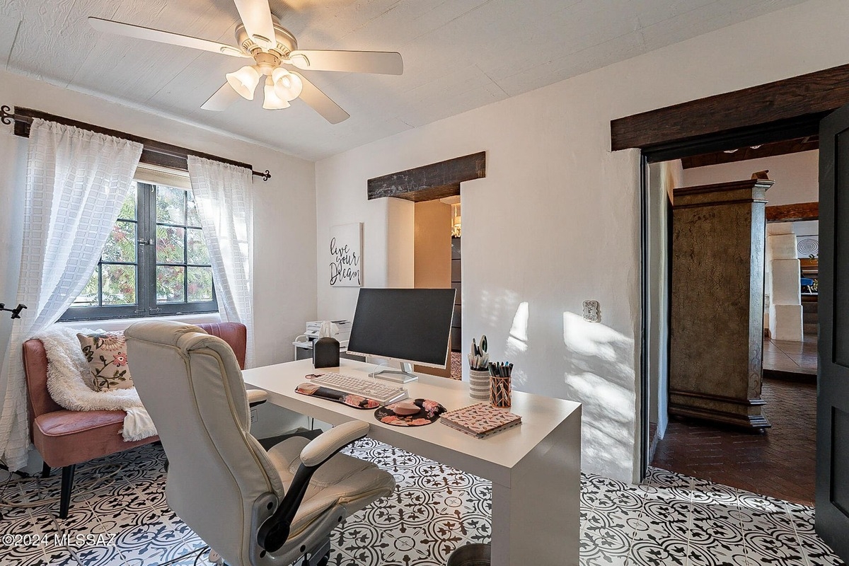 Home office features a white desk, computer, and chair, a pink couch, patterned flooring, and a ceiling fan, with a view of another room through an open doorway.