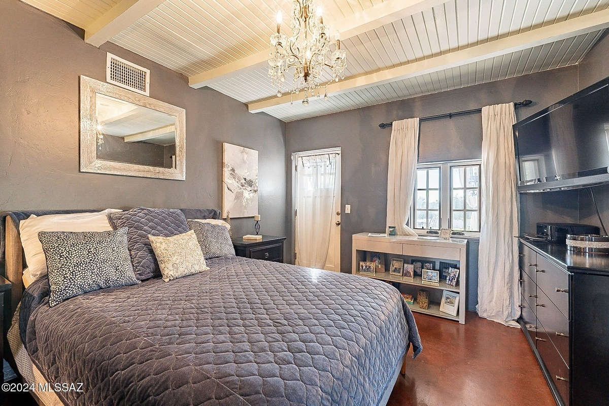 Bedroom features a gray quilted bedspread, a chandelier, gray walls, and a flat-screen TV mounted on a dark dresser, with a view of a door and window.