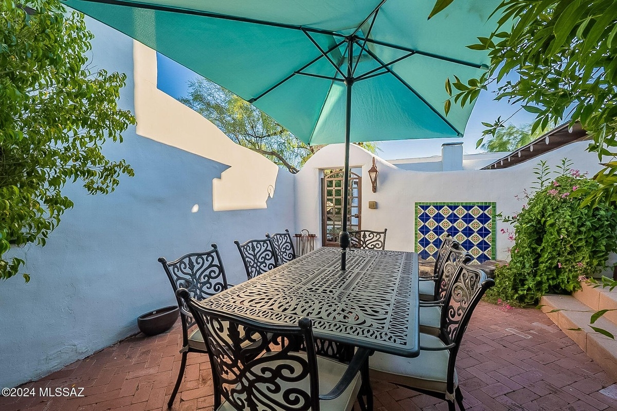 Ornate metal patio furniture sits under a teal umbrella in a courtyard with brick flooring and a tiled wall.