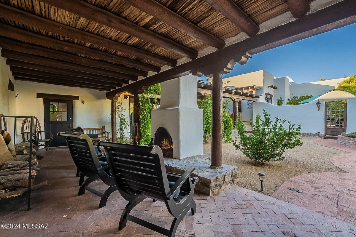 Covered patio with wooden beams, black chairs, and a view of a courtyard with a white stucco building and a fireplace.