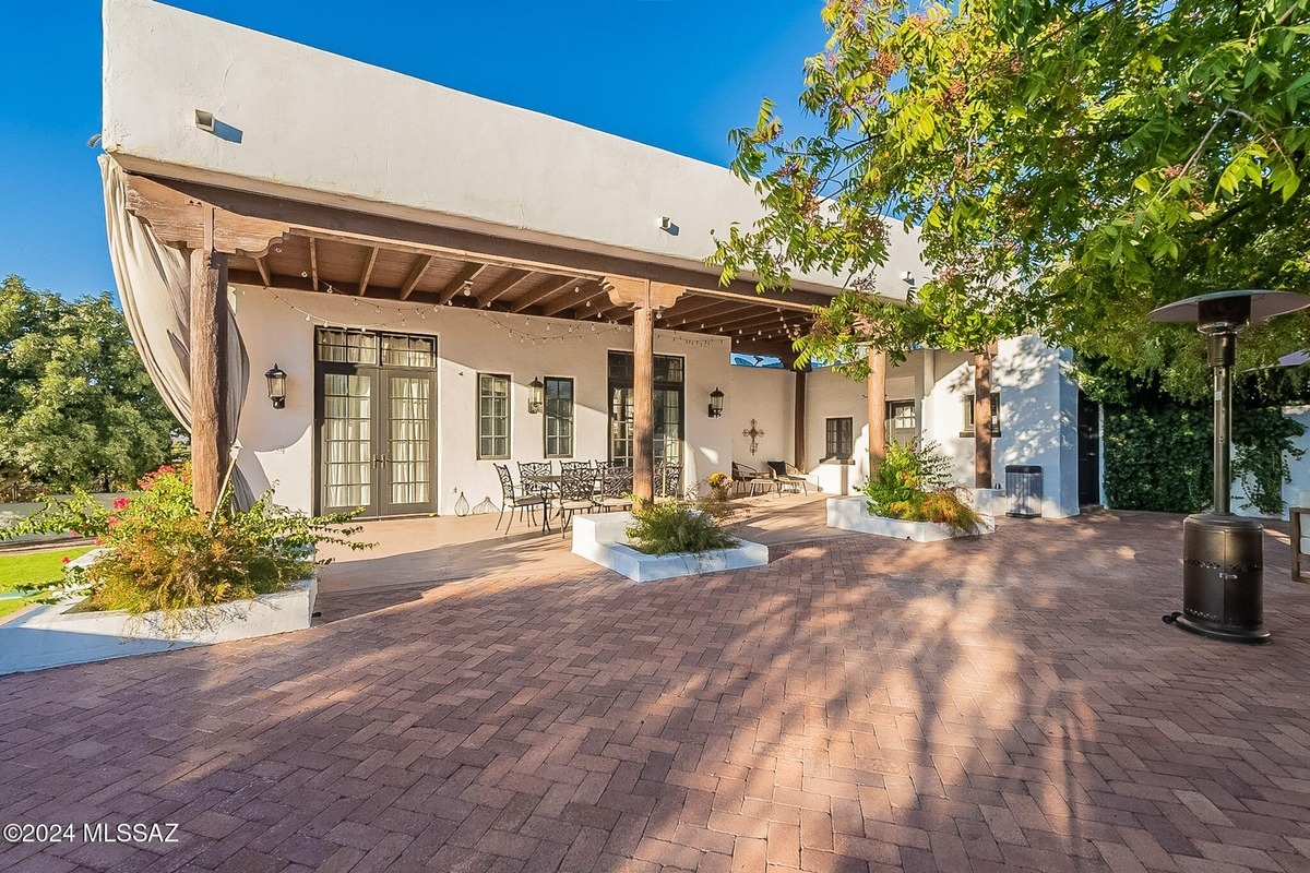 Outdoor patio area features a white stucco building with wooden beams, brick paving, and lush landscaping, with a patio heater in the foreground.