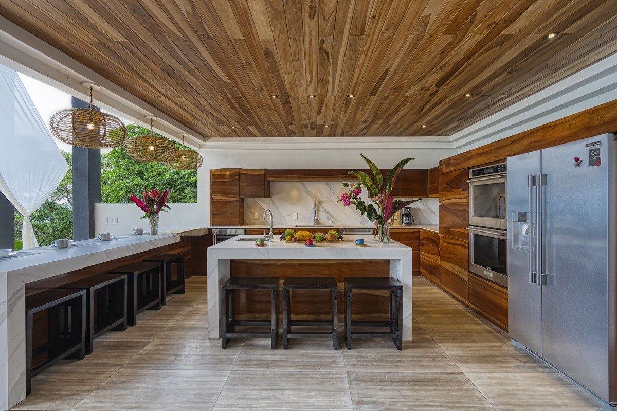 Outdoor kitchen has wood cabinets, marble countertops, and wood ceiling.