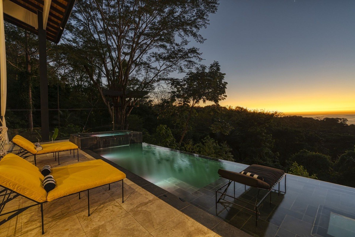 Infinity pool and lounge chairs overlook rainforest at sunset.