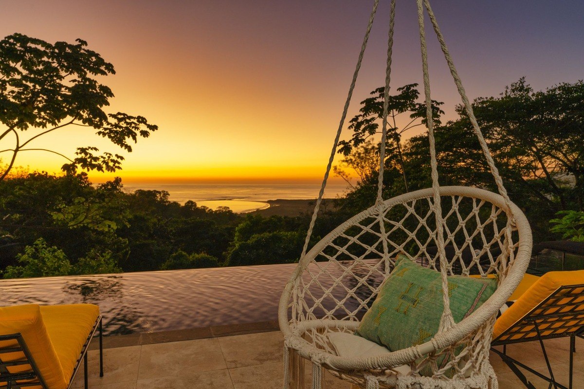 Hanging chair and lounge chairs overlook ocean at sunset.