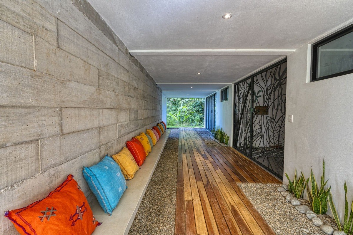 Covered walkway has colorful pillows on bench and wood flooring.