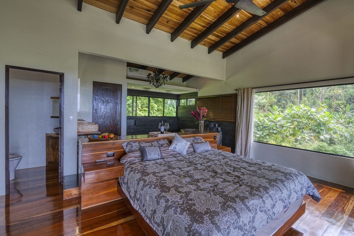 Master bedroom has large bed, wood floors, and forest view.