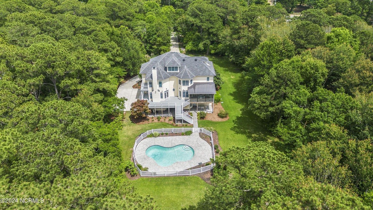 House has kidney-shaped pool, green lawn, and is surrounded by trees.