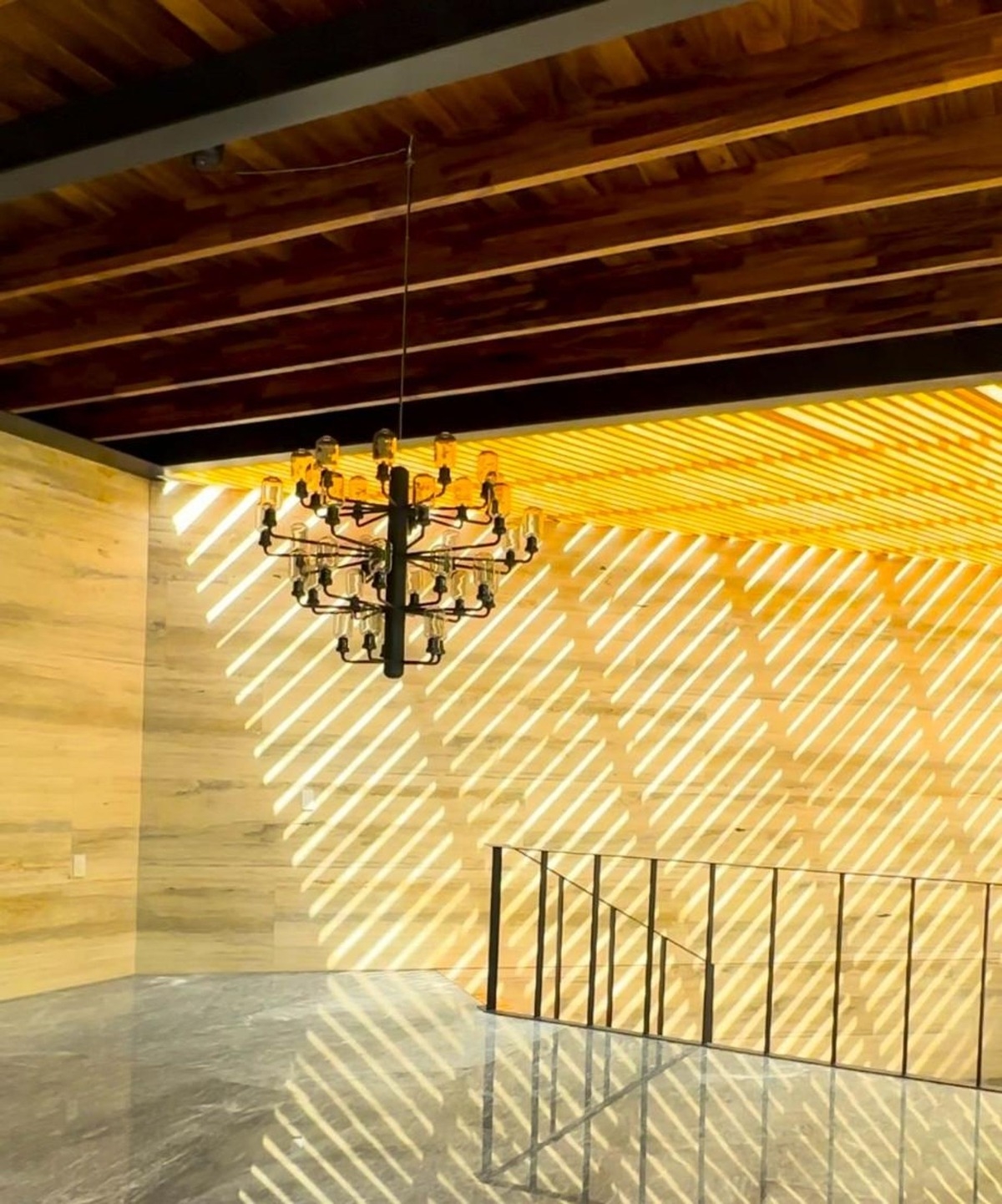 Black chandelier hangs from a wooden ceiling illuminated by diagonal light patterns.