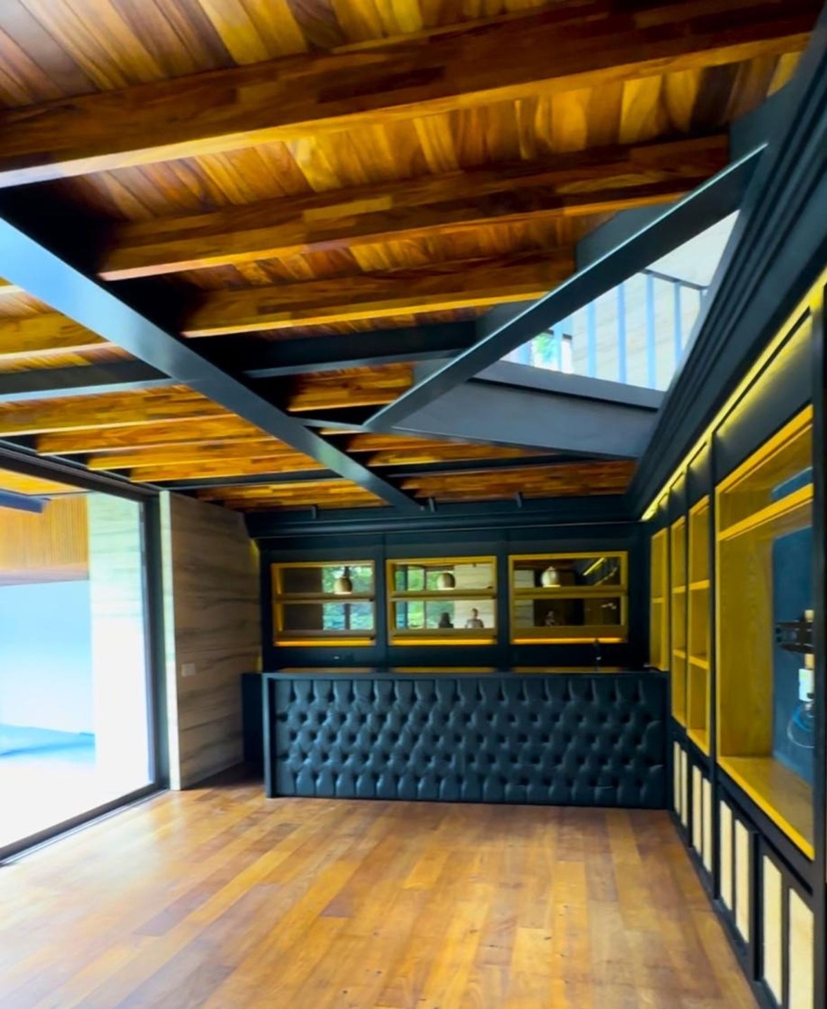 Room featuring a black tufted bar counter with golden shelving and wooden ceiling beams.