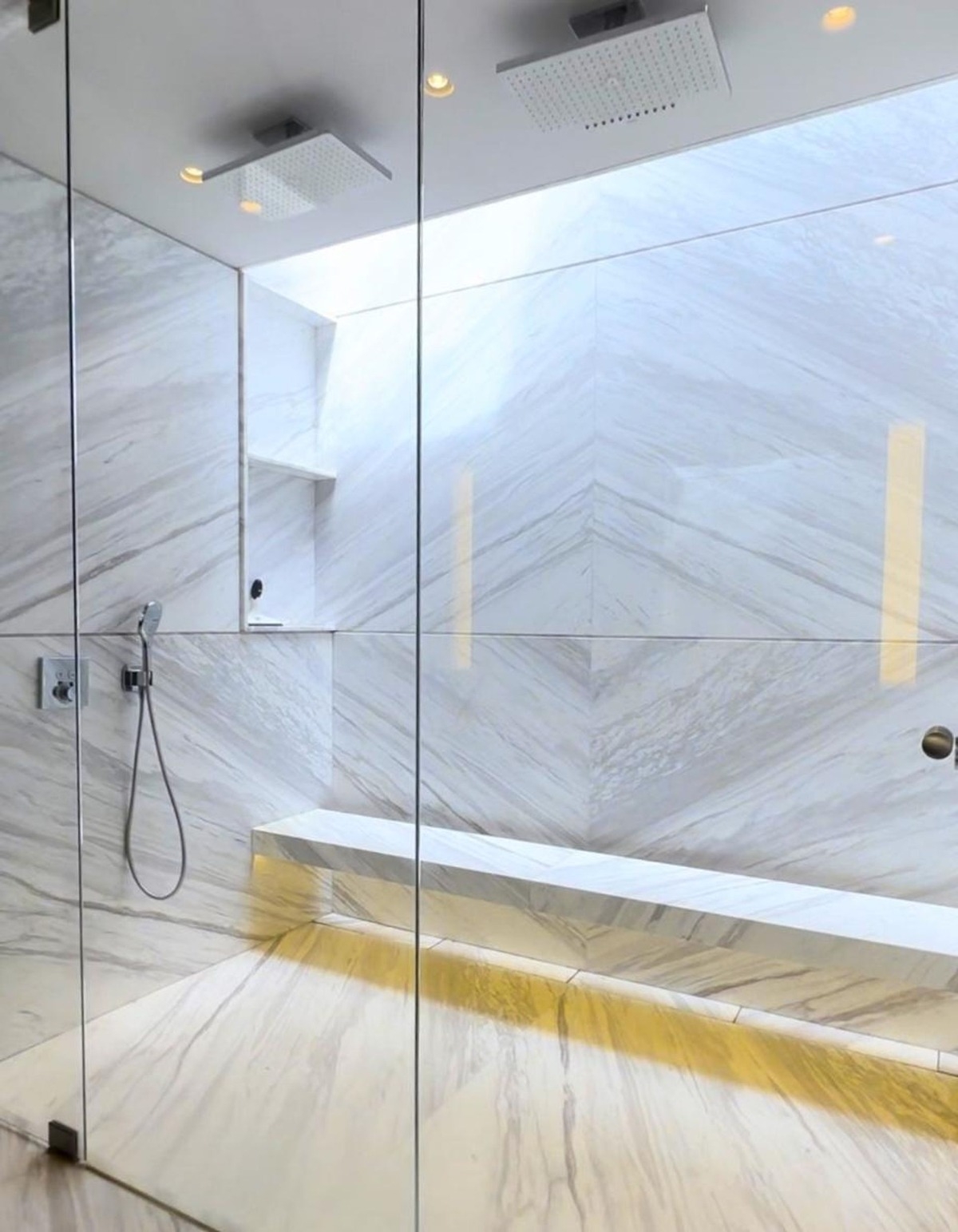 Glass-enclosed shower with marble walls and ceiling-mounted rain showers.