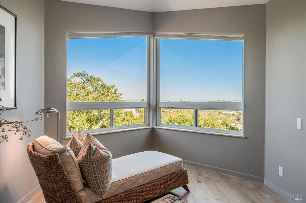 A chaise lounge placed near large corner windows with views of greenery and the horizon.