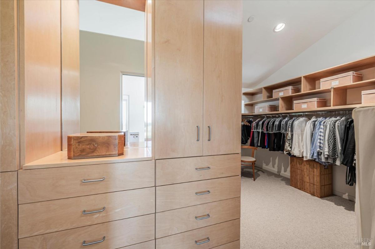 Spacious walk-in closet with a large mirror on one wall and is filled with built-in wooden shelves and drawers, and there are clothes hanging on racks.