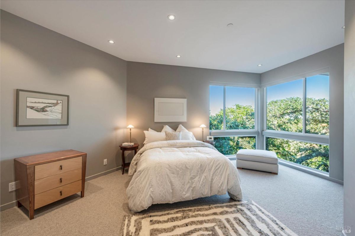 A bedroom with a large bed, minimal decor, and expansive windows overlooking trees.