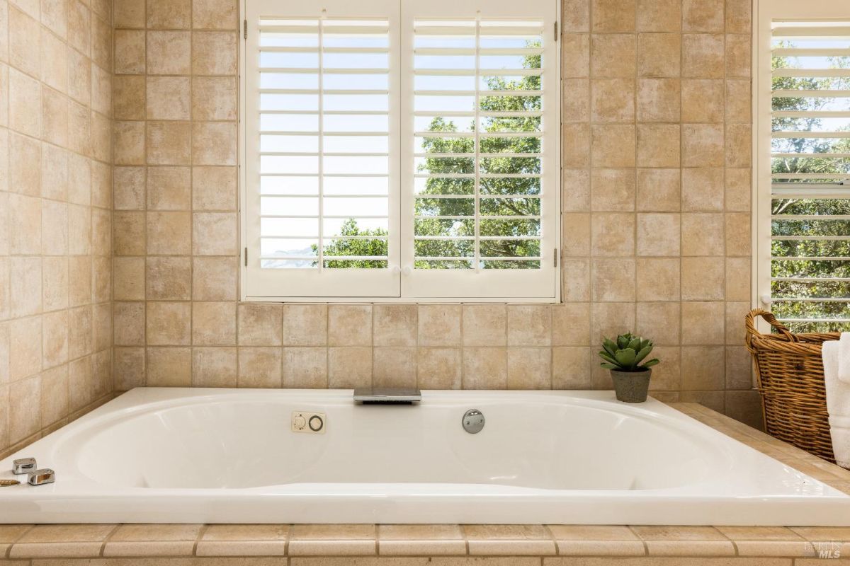 Bathroom with a tiled soaking tub beneath windows that provide views of trees.