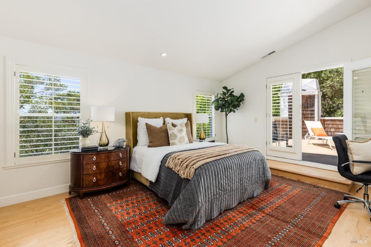 Bright bedroom with a green bedspread, red bedside tables, and views of greenery through windows and patio access.