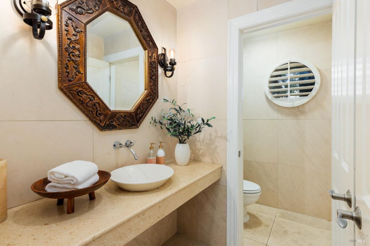 Elegant half-bathroom with a carved wood-framed mirror, light beige walls, and decorative lighting fixtures.