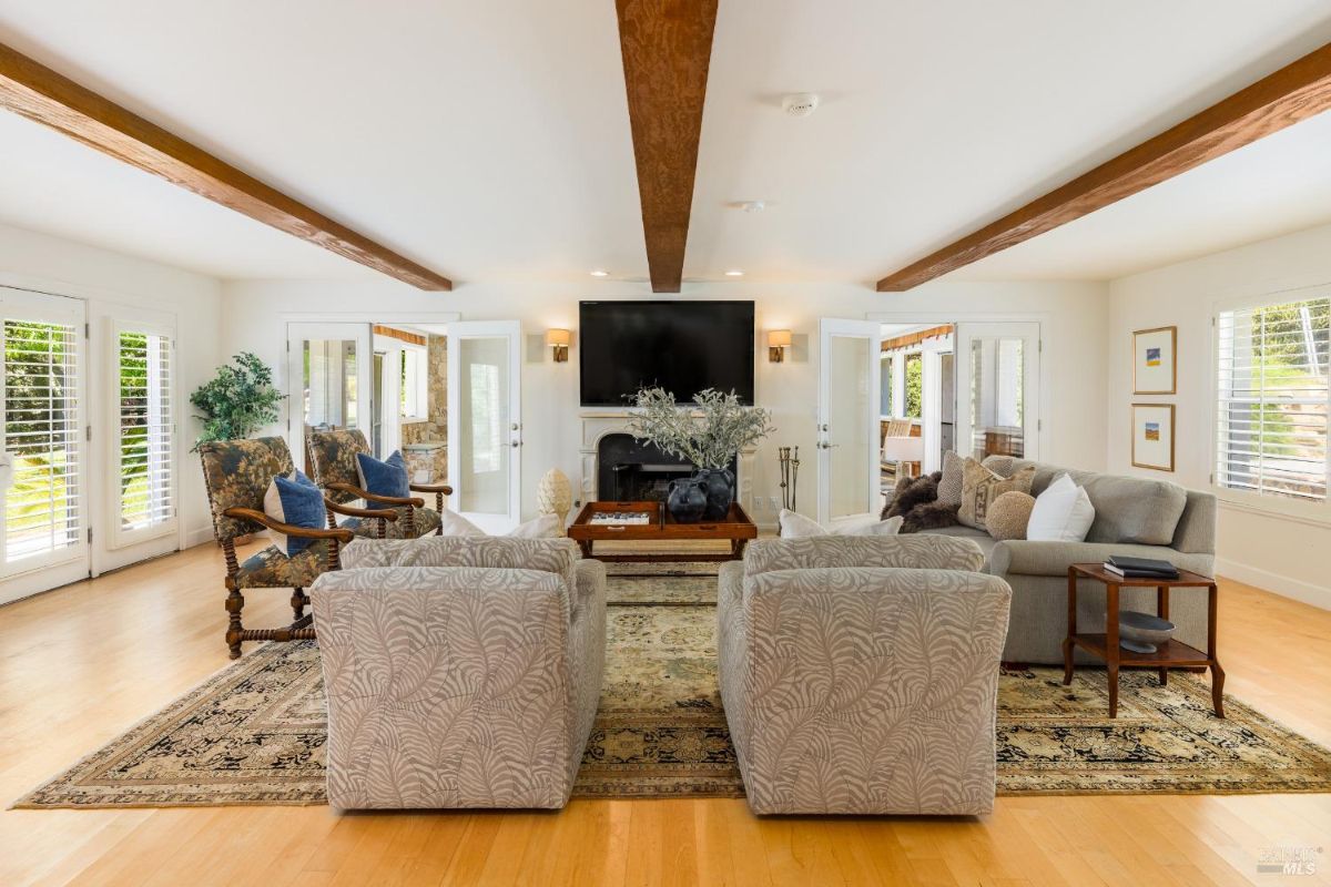 Living room with a large wall-mounted TV, exposed wooden beams, and light-colored furniture.