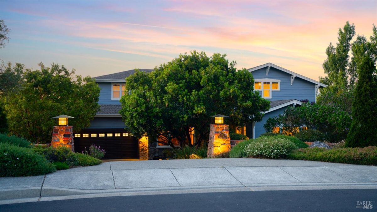 House exterior at sunset with a well-lit driveway, trees, and decorative stone pillars.