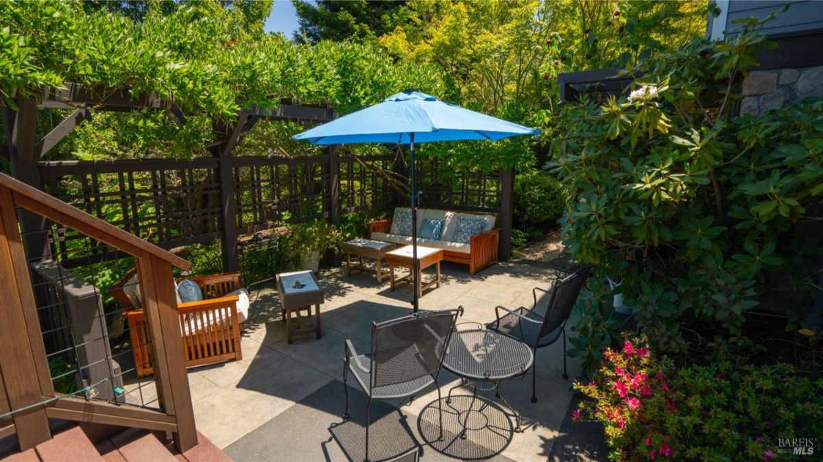 Cozy outdoor sitting area with metal chairs, wooden furniture, and vibrant plants in a shaded corner.