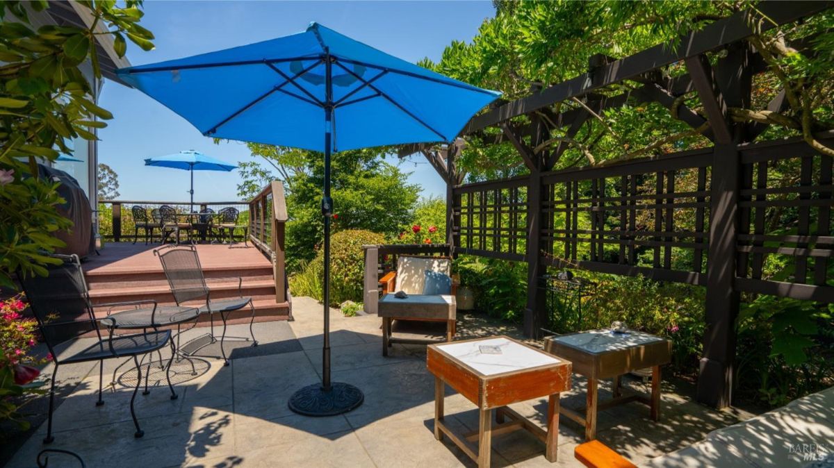 Outdoor patio with wooden furniture, blue umbrellas, and lattice structures surrounded by lush greenery.